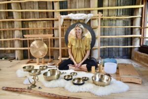 A woman sits cross-legged on a rug surrounded by various sound bowls, gongs, and sound healing instruments in a wooden room with rustic walls and natural wood beams. She is smiling and wearing a yellow top. - Fair Harvest Permaculture