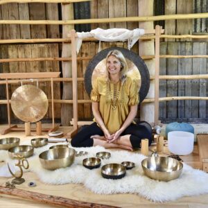 Ute Coleman sits cross-legged on a rug surrounded by various sound bowls, gongs, and sound healing instruments in a wooden room with rustic walls and natural wood beams. She is smiling and wearing a yellow top. - Fair Harvest Permaculture