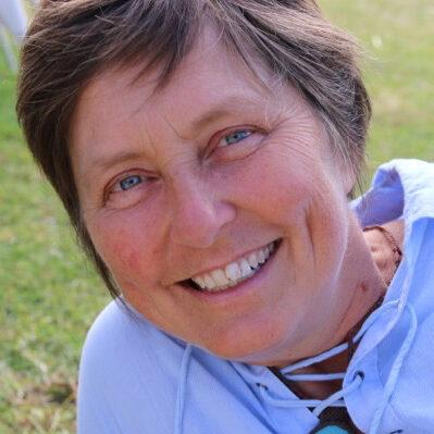 A woman with short brown hair and blue eyes is smiling at the camera while lying on the grass. She is wearing a light blue shirt and a necklace. The background is grassy with part of a white chair visible. - Fair Harvest Permaculture A woman with short brown hair and blue eyes is smiling at the camera while lying on the grass. She is wearing a light blue shirt and a necklace. The background is grassy with part of a white chair visible. - Fair Harvest Permaculture