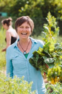 A woman stands outdoors in a garden, smiling and holding a bunch of leafy greens. She is wearing a light blue shirt and a necklace. Another person is blurred in the background among green plants. - Fair Harvest Permaculture