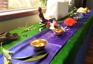 A table covered with a green cloth and a purple runner displays bowls of water, flowers, a small Buddha statue, fruit, leaves, and decorative objects near a window with sunlight coming in. - Fair Harvest Permaculture