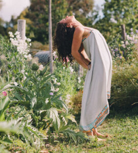 A person in a long, light-coloured dress stands outdoors in a garden, leaning back with hands on their lower back and head tilted up. There are green plants and flowers around, and sunlight is visible. - Fair Harvest Permaculture