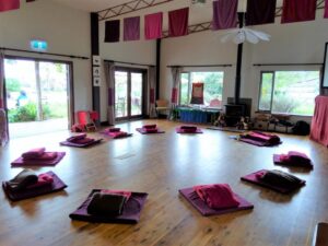 A spacious room with wooden floors has meditation cushions arranged in a circle. Folded blankets are placed on each cushion. Large windows let in natural light, and colourful fabric panels hang from the ceiling. - Fair Harvest Permaculture