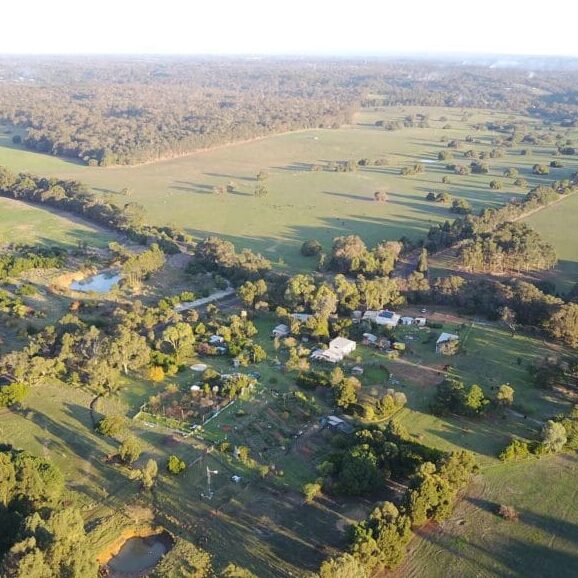 Aerial view of a rural landscape with scattered buildings, gardens, trees, ponds, and open green fields stretching into the distance under a clear sky. - Fair Harvest Permaculture Aerial view of a rural landscape with scattered buildings, gardens, trees, ponds, and open green fields stretching into the distance under a clear sky. - Fair Harvest Permaculture